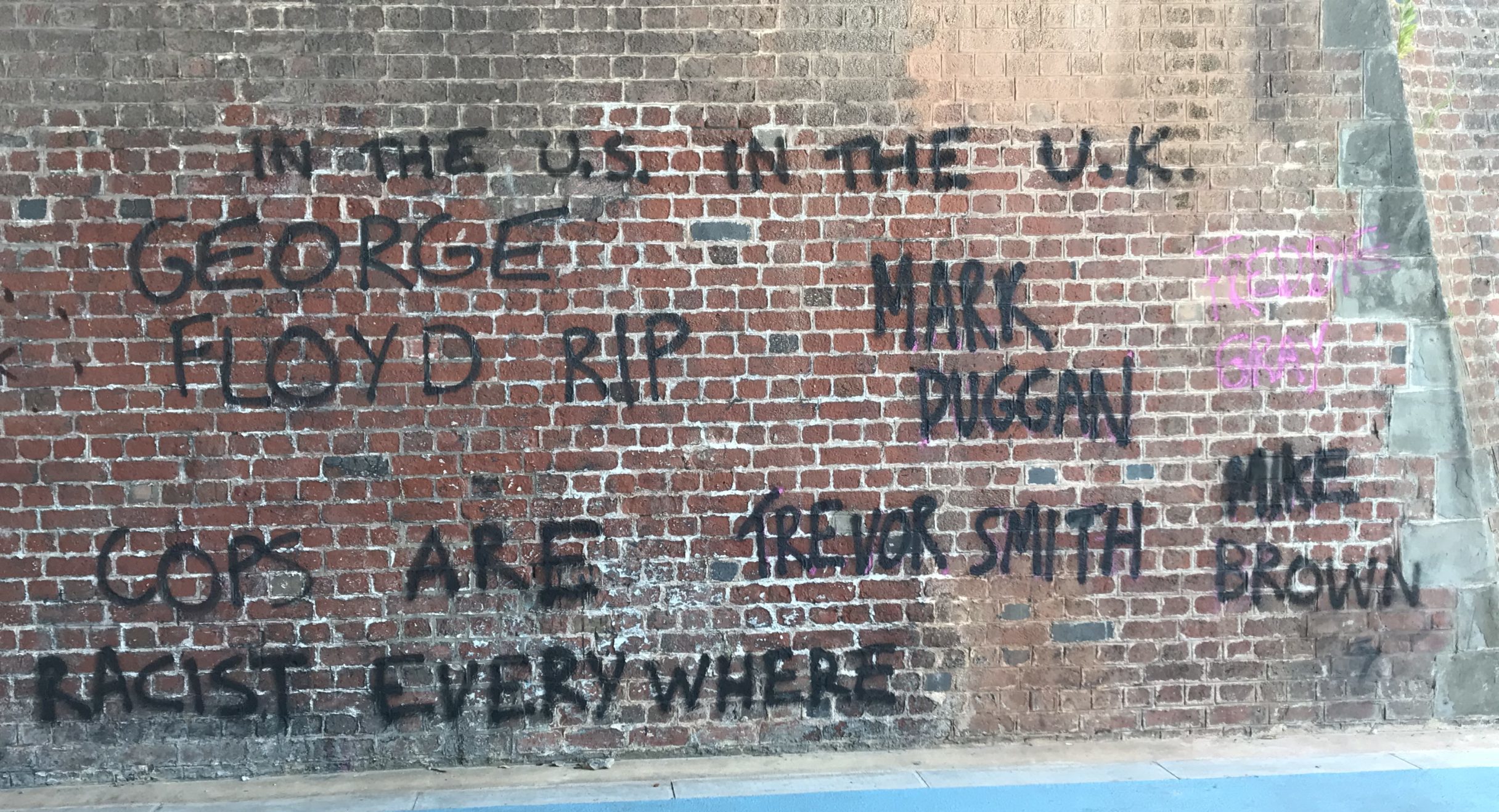 A brick wall in Bristol. On the wall is some graffiti rendered in black spray paint: "In the US, in the UK, the police are racist everywhere: George Floyd RIP. Mark Duggan. Trevor Smith. Mike Brown"